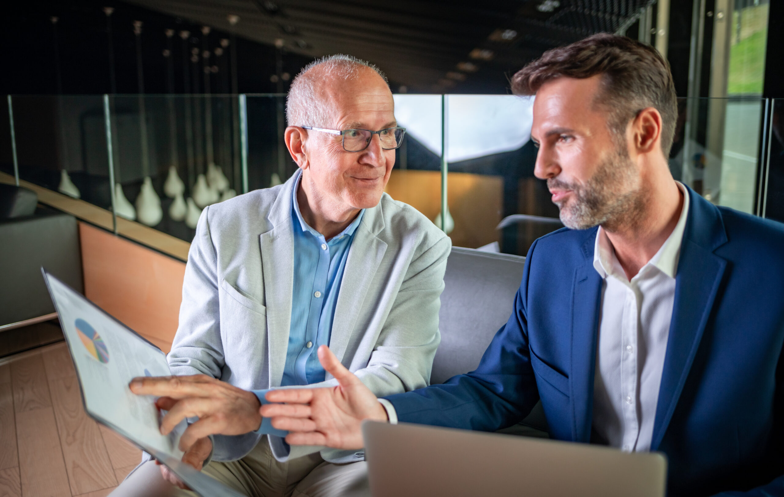 Younger and older businessman talking about documents during meeting in modern business lounge Financial Advisor explaining the two advantages of obtaining a signed HIPAA with every life insurance aplication