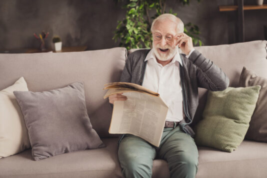 Photo of smiling pensioner wear grey cardigan sitting couch reading newspaper arm glasses indoors house flat Newspaper reader is delighted with news you can use in the Wall Street Journal