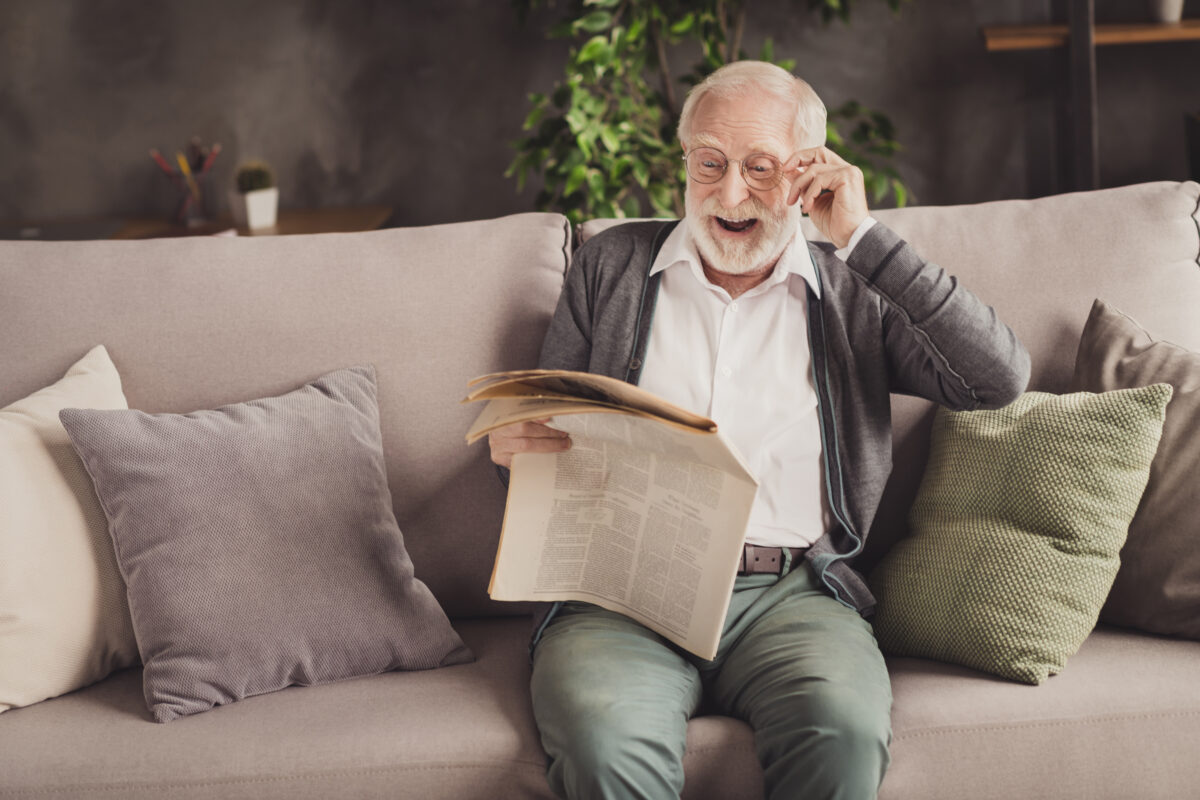 Photo of smiling pensioner wear grey cardigan sitting couch reading newspaper arm glasses indoors house flat Newspaper reader is delighted with news you can use in the Wall Street Journal