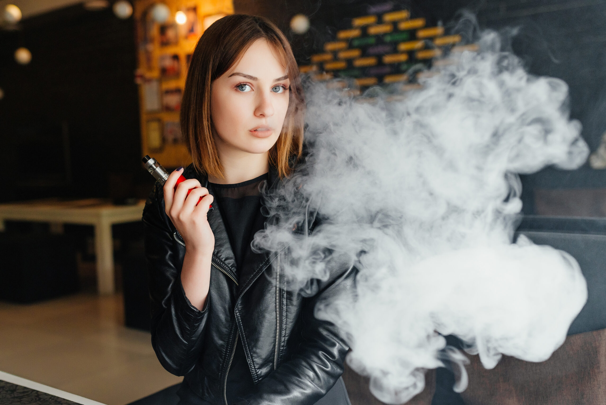 Vaping. Young beautiful girl smoking ( vaping ) e-cigarette with smoke in restaurant. Vapor concept. An attractive woman is vaping with a smile because she learned about Underwriting Exceptions for Tobacco Users Vaper Kayla
